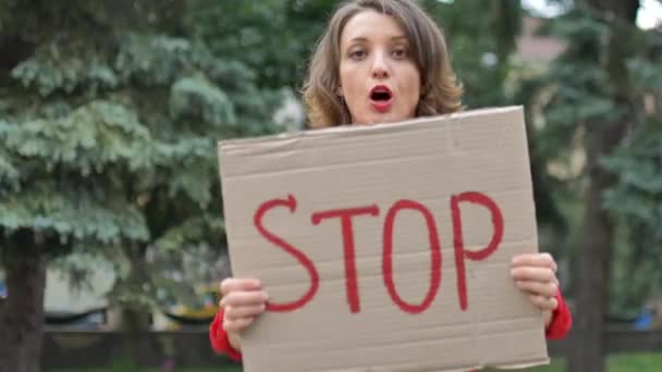 Молода жінка, що протестує в червоній сорочці, тримає знак протесту на дошці з гаслом Стоп для публічної демонстрації на фоні дерев . — стокове відео