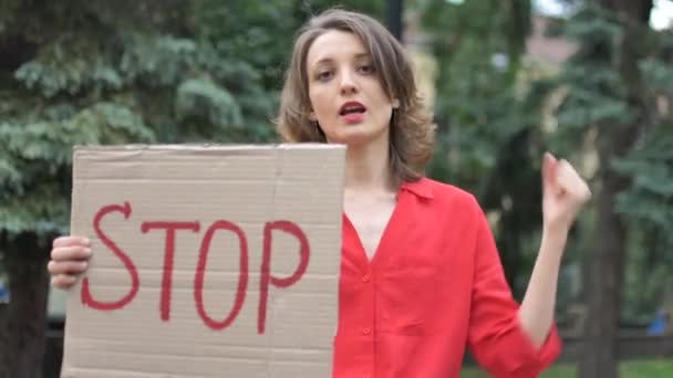 Jonge protesterende vrouw in rood shirt houdt protestbord plakbord met slogan Stop voor publieke demonstratie op bomen achtergrond. — Stockvideo