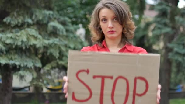 Jonge protesterende vrouw in rood shirt houdt protestbord plakbord met slogan Stop voor publieke demonstratie op bomen achtergrond. — Stockvideo