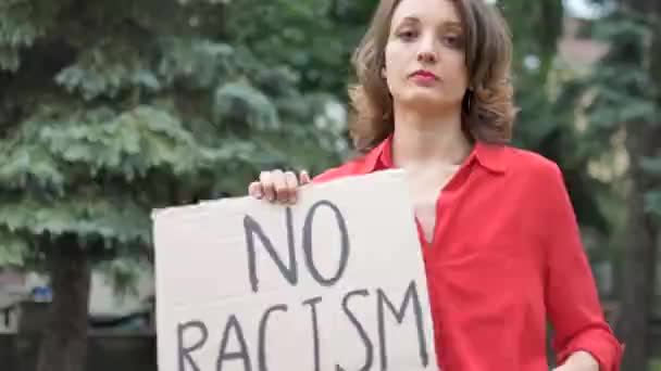 Sconvolto giovane donna protestante in camicia rossa tiene segno di protesta cartello foglio largo con slogan Nessun razzismo per la manifestazione pubblica su sfondo alberi. — Video Stock