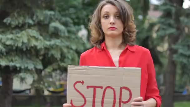 Jonge protesterende vrouw in rood shirt houdt protestbord plakbord met slogan Stop voor publieke demonstratie op bomen achtergrond. — Stockvideo