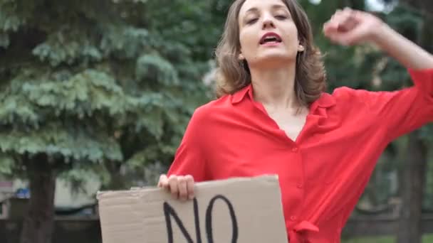 Upset young protesting woman in red shirt holds protest sign broadsheet placard with slogan No racism for public demonstration on trees background. — Stock Video