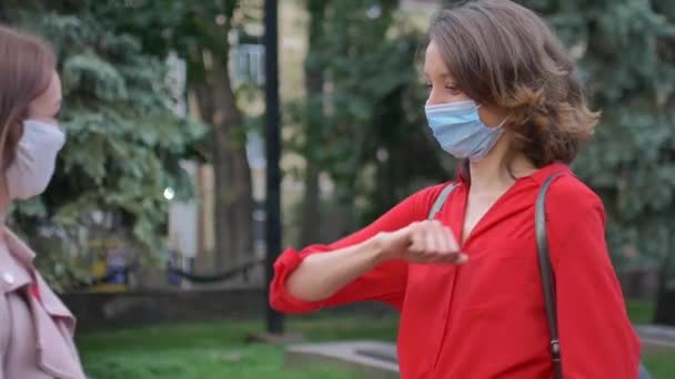 Happy meeting of two female friends wearing face mask and keep social distance at the street on trees background, friendship and communication during quarantine — Stock Video