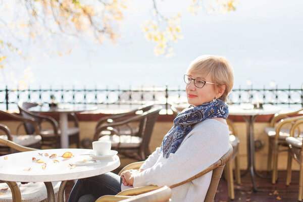 Relaxed senior woman is sitting and resting in autumn street cafe outside. Beautiful retired female person is enjoying life and coffee. Concept of life satisfaction, calm, wellbeing.