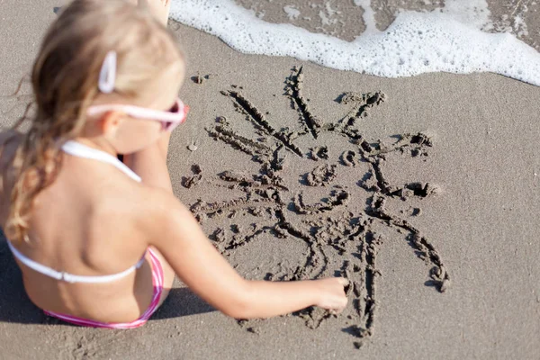 Kid is drawing smiling sun on sand at beach. Concept of children picture, sand painting at summer vacation, holiday and travel. Little girl is sitting in waves outdoors. Child is enjoying summertime.