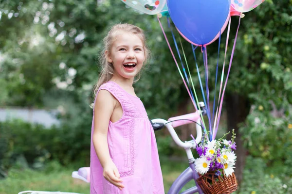 Little girl is surprised of Happy Birthday gift. Kid gets bicycle with balloons, basket with bouquet of summer flowers. Child is smiling, having fun, laughing. Lifestyle moment, real candid emotions.