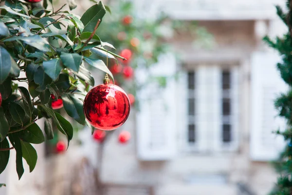 Red Christmas balls and ornaments on branches of tree. Background with stone house, white window, wooden shutters
