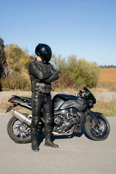 O motociclista de preto usando um capacete olha para uma motocicleta — Fotografia de Stock