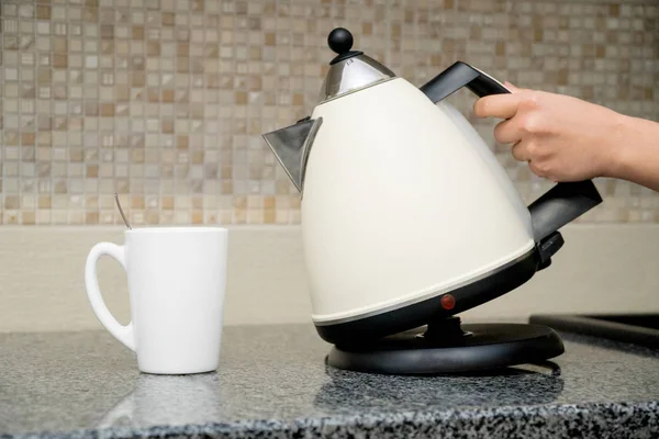 La mano delle donne tiene bollitore elettrico di vetro su una cucina domestica — Foto Stock