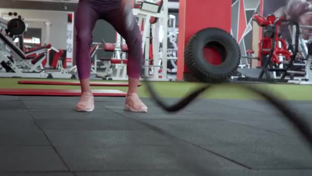Молодая девушка упорно тренируется с боевой веревкой во время кросс-тренировки. Красивая девушка в спортивном клубе делает различные виды упражнений на тренажерах — стоковое видео