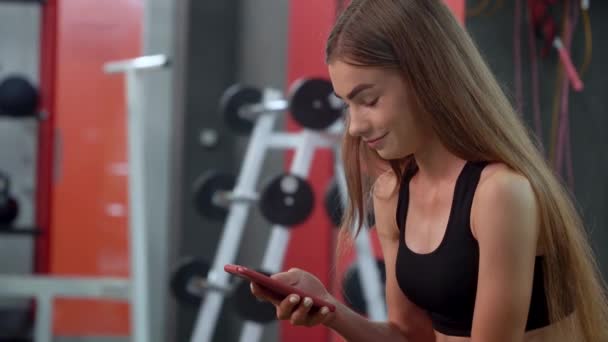 Chica joven sonrisa y utiliza el teléfono inteligente mientras descansa en el banco después de su entrenamiento intensivo — Vídeo de stock