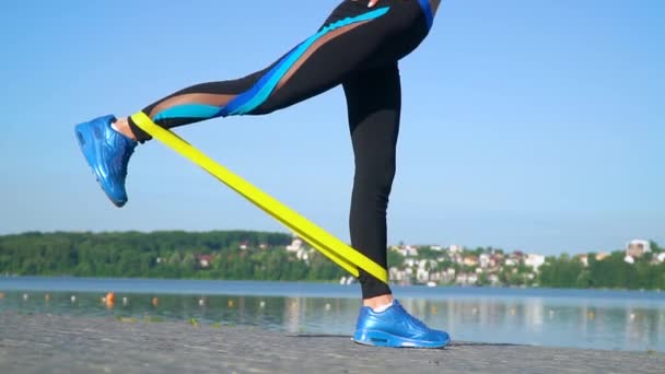 Ajuste mujer atlética en ropa deportiva usando banda elástica con piernas estiradas y todo el cuerpo cerca del lago. Chica joven haciendo ejercicios matutinos en el parque, actividad física, bienestar — Vídeo de stock