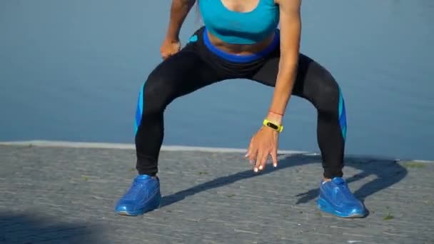 Young woman, dressed in sports suit, does a split squat on a lake coast in summer — Stock Video