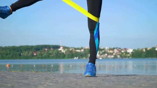 Fit athletische Frau in Sportbekleidung mit Gummiband mit Stretchbeinen und Ganzkörper in der Nähe des Sees. junges Mädchen macht Morgengymnastik im Park, Fitness-Aktivität, Wellness — Stockvideo
