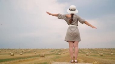 Şapkalı genç kadın, yaz akşamı balyalarla buğday tarlasının ortasında elleriyle gökyüzüne bakıyor.