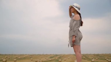 Şapkalı genç kadın yaz akşamı balyalarla buğday tarlasının gökyüzüne bakıyor.