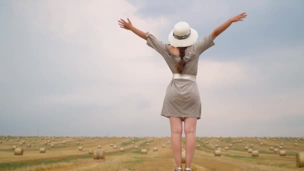 Mooie jonge vrouw in een hoed kijkt in de lucht met haar handen naar de hemel in het midden van een tarwe veld met Balen in de zomeravond — Stockvideo