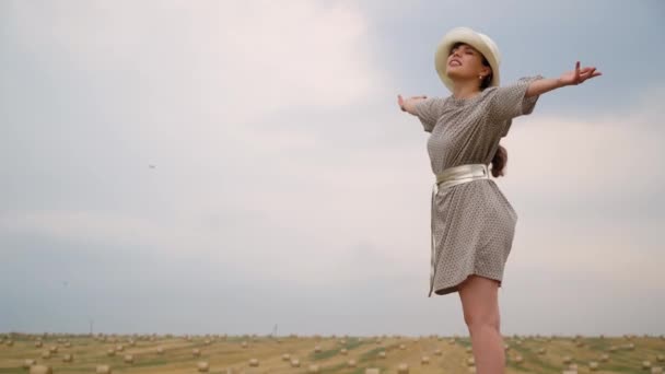 Hermosa joven en un sombrero mira al cielo con las manos hacia el cielo en medio de un campo de trigo con fardos en la noche de verano — Vídeo de stock