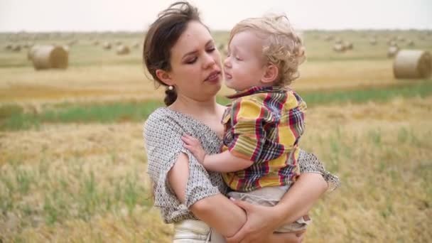 Mulher bonita com o filho pequeno fair-haired em seus braços no meio de um campo de trigo com fardos na noite do verão. Mãe beija seu filho — Vídeo de Stock