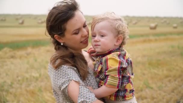 Mulher bonita com o filho pequeno fair-haired em seus braços no meio de um campo de trigo com fardos na noite do verão. Mãe beija seu filho — Vídeo de Stock