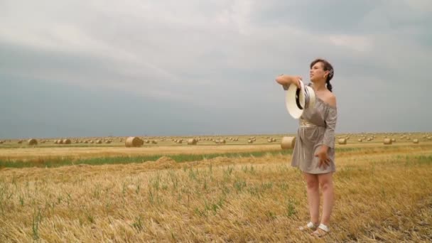 Young woman throws her hat on the ground of a wheat field with bales in summer evening — Stock Video