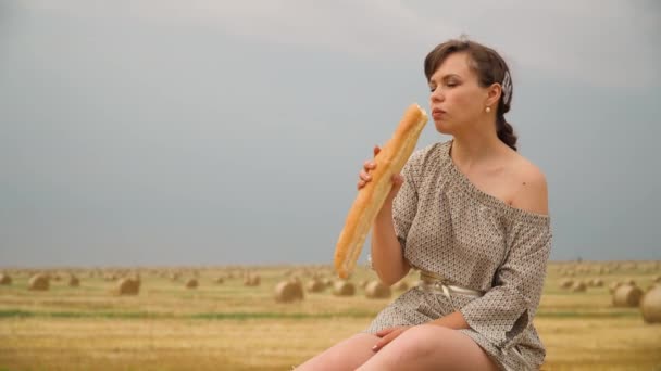 Jovem sentada em cima do fardo e comendo pão no meio de um campo de trigo com fardos na noite de verão — Vídeo de Stock