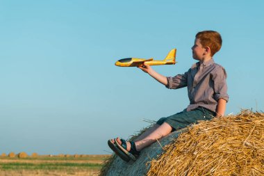Kızıl saçlı çocuk balyalarla buğday tarlasında oyuncak bir uçakla oynuyor.