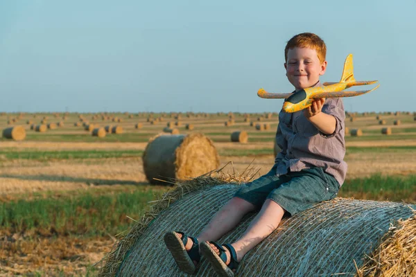 O menino ruivo brinca com um avião de brinquedo em um campo de trigo com fardos — Fotografia de Stock