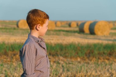 Kırmızı saçlı bir çocuk saman balyaları bir arka plan üzerinde bir buğday tarlasında yan duruyor