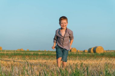 Kızıl saçlı çocuk saman balyalarının arka planında buğday tarlasının önüne geçiyor.