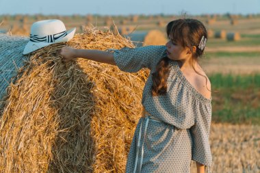 Saman balyaları yakınındaki bir buğday tarlasında poz veren uzun saçlı ve şapkalı genç güzel kadın