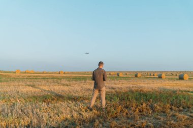 Genç bir adam balyalarla buğday tarlasında duruyor ve insansız hava aracını kontrol ediyor.