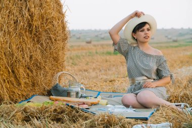 Güzel genç kadın yaz akşamı bir buğday tarlasının ortasında bir saman balyası yakınında piknik yapıyor