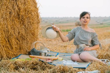 Güzel genç kadın yaz akşamı bir buğday tarlasının ortasında bir saman balyası yakınında piknik yapıyor