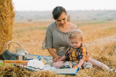 Yaz akşamı buğday tarlasının ortasında saman balyası yakınında piknik küçük sarışın oğlu ile güzel genç kadın