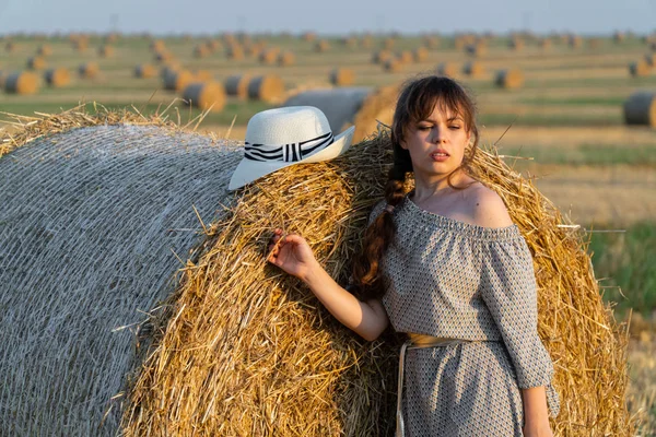 Saman balyaları yakınındaki bir buğday tarlasında poz veren uzun saçlı ve şapkalı güzel genç kız