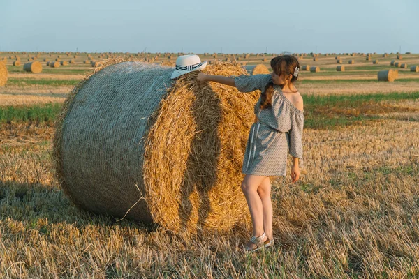 Beautiful young girl with long hair and hat posing on a wheat field near hay bales — Stock Photo, Image