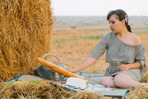 Mulher bonita fazendo um piquenique perto de um fardo de feno no meio de um campo de trigo na noite de verão — Fotografia de Stock