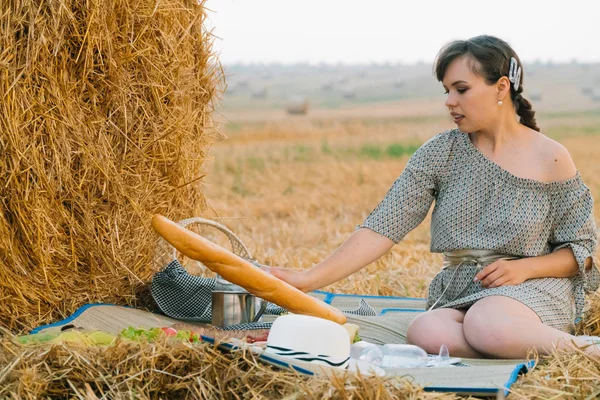 Menina bonita fazendo um piquenique perto de um fardo de feno no meio de um campo de trigo na noite de verão — Fotografia de Stock