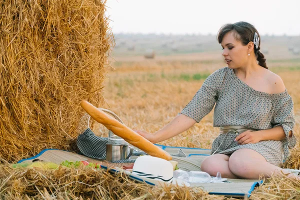 Mulher bonita fazendo um piquenique perto de um fardo de feno no meio de um campo de trigo na noite de verão — Fotografia de Stock