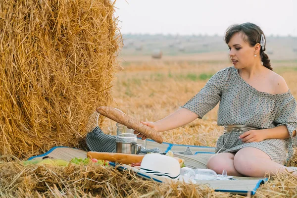 Menina bonita fazendo um piquenique perto de um fardo de feno no meio de um campo de trigo na noite de verão — Fotografia de Stock