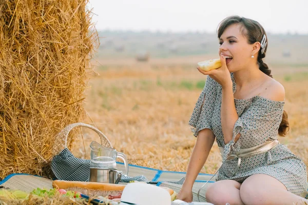 Прекрасная молодая женщина устраивает пикник и ест хлеб возле тюка сена посреди пшеничного поля летним вечером — стоковое фото