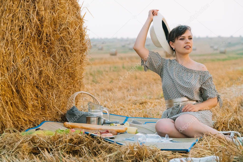 Beautiful young woman having a picnic near a hay bale in the middle of a wheat field in summer evening