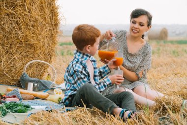 Yaz akşamı buğday tarlasının ortasında saman balyası yakınında piknikte kızıl saçlı oğlu olan ve havuç suyu içen güzel genç kadın