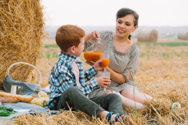 Yaz akşamı buğday tarlasının ortasında saman balyası yakınında piknikte kızıl saçlı oğlu olan ve havuç suyu içen güzel genç kadın