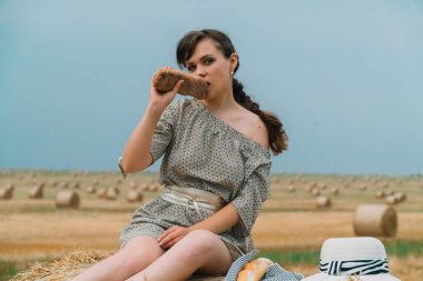 Genç kız yaz akşamı balya ile bir buğday tarlasının ortasında balya üzerinde oturan ve ekmek yemek