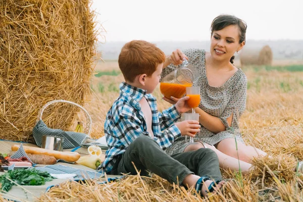 Yaz akşamı buğday tarlasının ortasında saman balyası yakınında piknikte kızıl saçlı oğlu olan ve havuç suyu içen güzel genç kadın