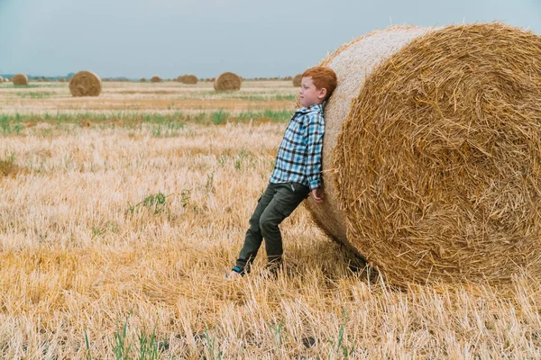 O menino ruivo apoiou-se em um fardo de palha no meio de um campo de trigo com fardos em uma noite de verão. — Fotografia de Stock