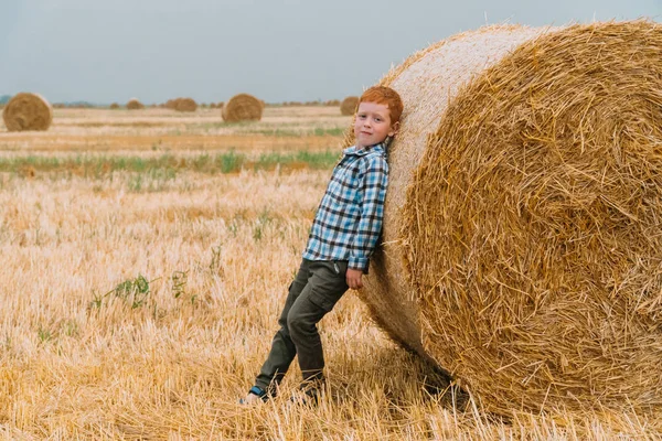 O menino ruivo apoiou-se em um fardo de palha no meio de um campo de trigo com fardos em uma noite de verão. — Fotografia de Stock