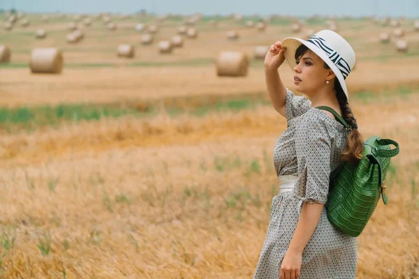 Mulher bonita em um chapéu e com uma mochila no meio de um campo de trigo com fardos em uma noite de verão — Fotografia de Stock